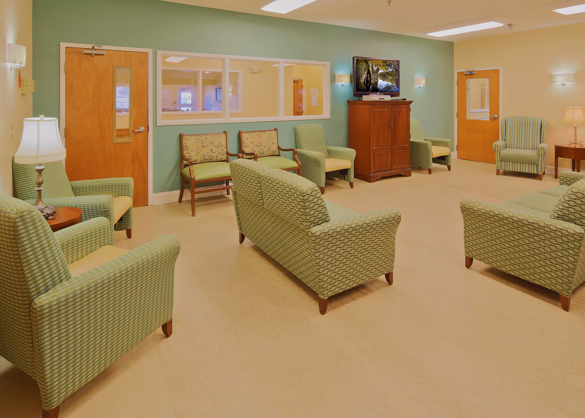 A spacious and well-lit living room in a memory care facility with multiple green patterned armchairs and sofas arranged around the room. There are wooden doors with windows, a wooden cabinet with a TV on top, and soft wall lighting. The walls are painted green and beige, creating a calm and welcoming atmosphere.
