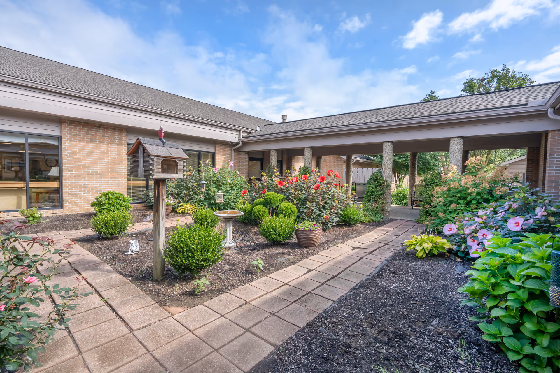 Outdoor garden area at Brookdale Mount Vernon featuring a paved walkway, various green shrubs, flowering plants including red and pink flowers, a birdhouse on a wooden post, and a birdbath. The garden is surrounded by a brick building with large windows and a covered walkway supported by stone columns under a partly cloudy sky.