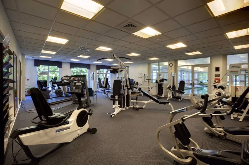A spacious fitness room with various exercise equipment including treadmills, stationary bikes, weight machines, and benches. The room has large windows letting in natural light and a carpeted floor. The ceiling has recessed lighting and there are mirrors on the walls.