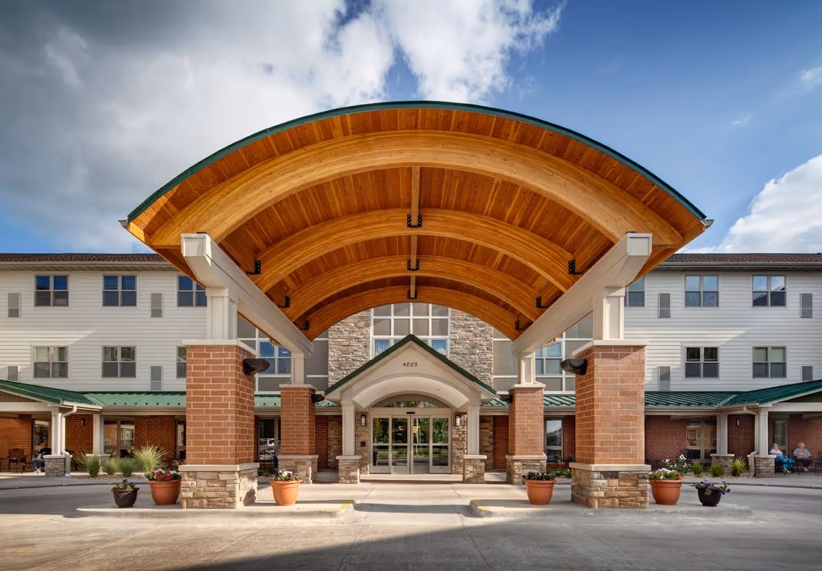 Front entrance of a multi-story senior living building with a large curved wooden canopy supported by brick columns and potted plants.