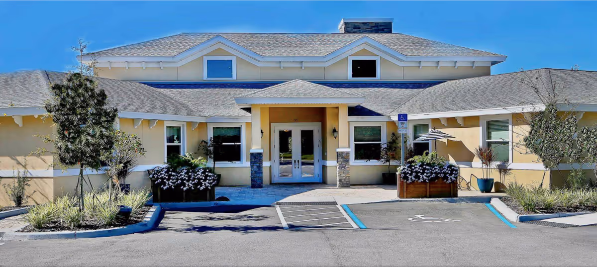 Front exterior view of a single-story building with a beige facade, multiple windows, a central entrance with double glass doors under a small covered porch, and a wheelchair accessible parking space in front. There are landscaped areas with small trees and flowering plants on either side of the entrance.