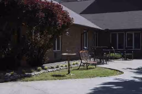 Outdoor courtyard area with a curved paved walkway, a water fountain, metal benches, and a table with chairs. The area is adjacent to a brick building with multiple windows and a sloped roof. There are bushes and grass surrounding the walkway.