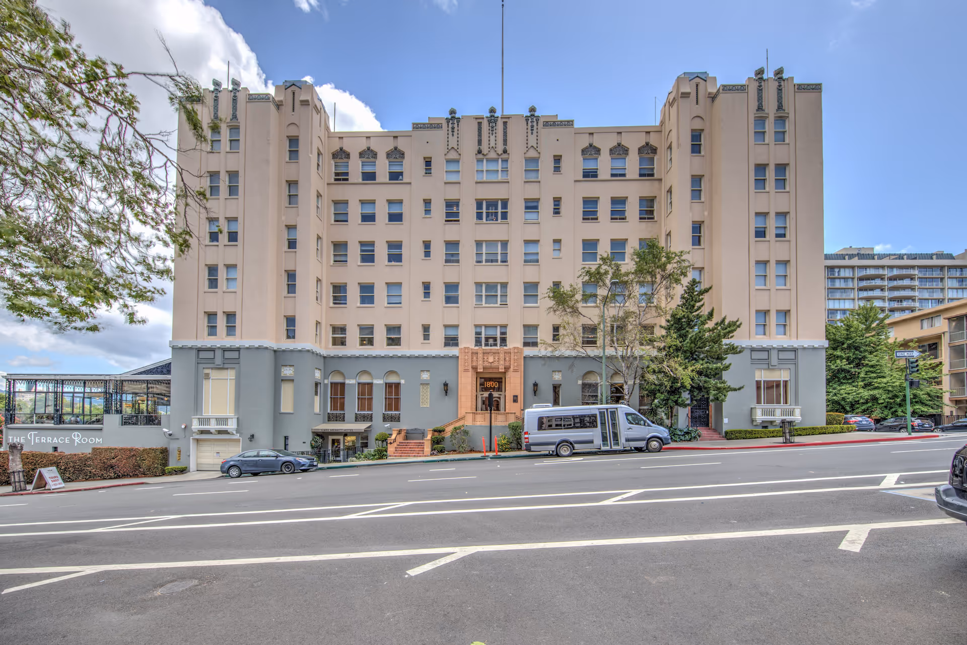 Exterior front view of a multi-story beige and gray building with several windows, a main entrance with steps, a van parked in front, and trees on either side. A sign on the left side reads 'The Terrace Room'. The sky is partly cloudy.