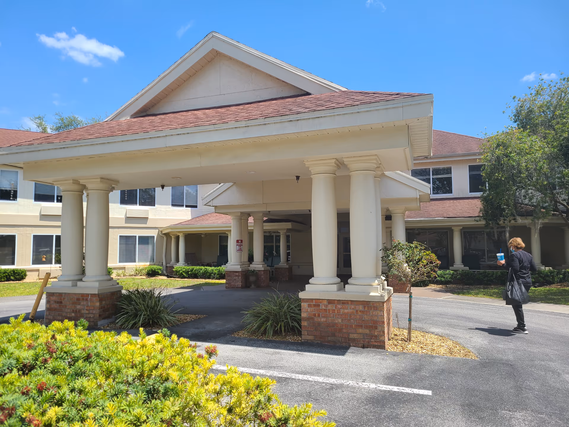 Exterior view of The Springs at Lady Lake Assisted Living facility showing the entrance with a covered driveway supported by large white columns with brick bases. The building has beige walls and a red roof. A person wearing a mask and holding a blue drink cup is walking near the entrance. Green shrubs and trees surround the driveway under a clear blue sky.