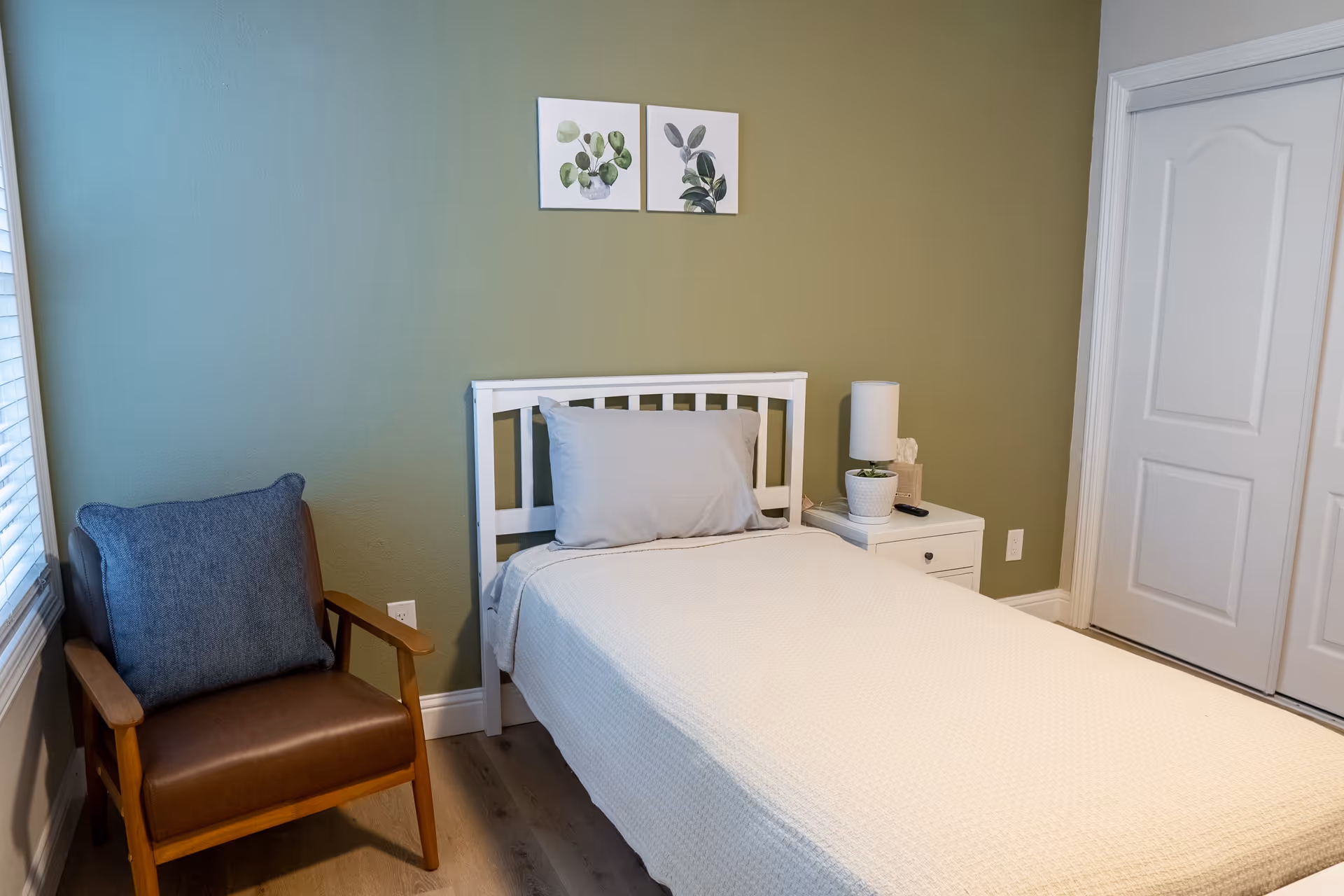 A simple bedroom with a single white bed covered with a cream-colored blanket and a gray pillow. Next to the bed is a white nightstand with a white lamp, a small potted plant, and a tissue box. To the left of the bed is a wooden armchair with a brown seat and a blue cushion. The walls are painted green, and two small framed botanical prints hang above the bed. There is a window with white blinds on the left side and a white closet door on the right.