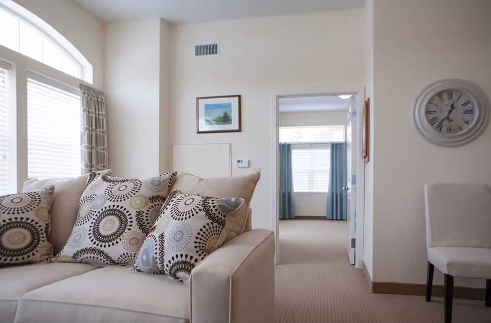 A cozy living room area with a beige sofa adorned with patterned cushions. There is a large window with blinds and curtains on the left side, a framed picture on the wall, and a doorway leading to another room with blue curtains and a window. A white chair and a round wall clock are visible on the right side.