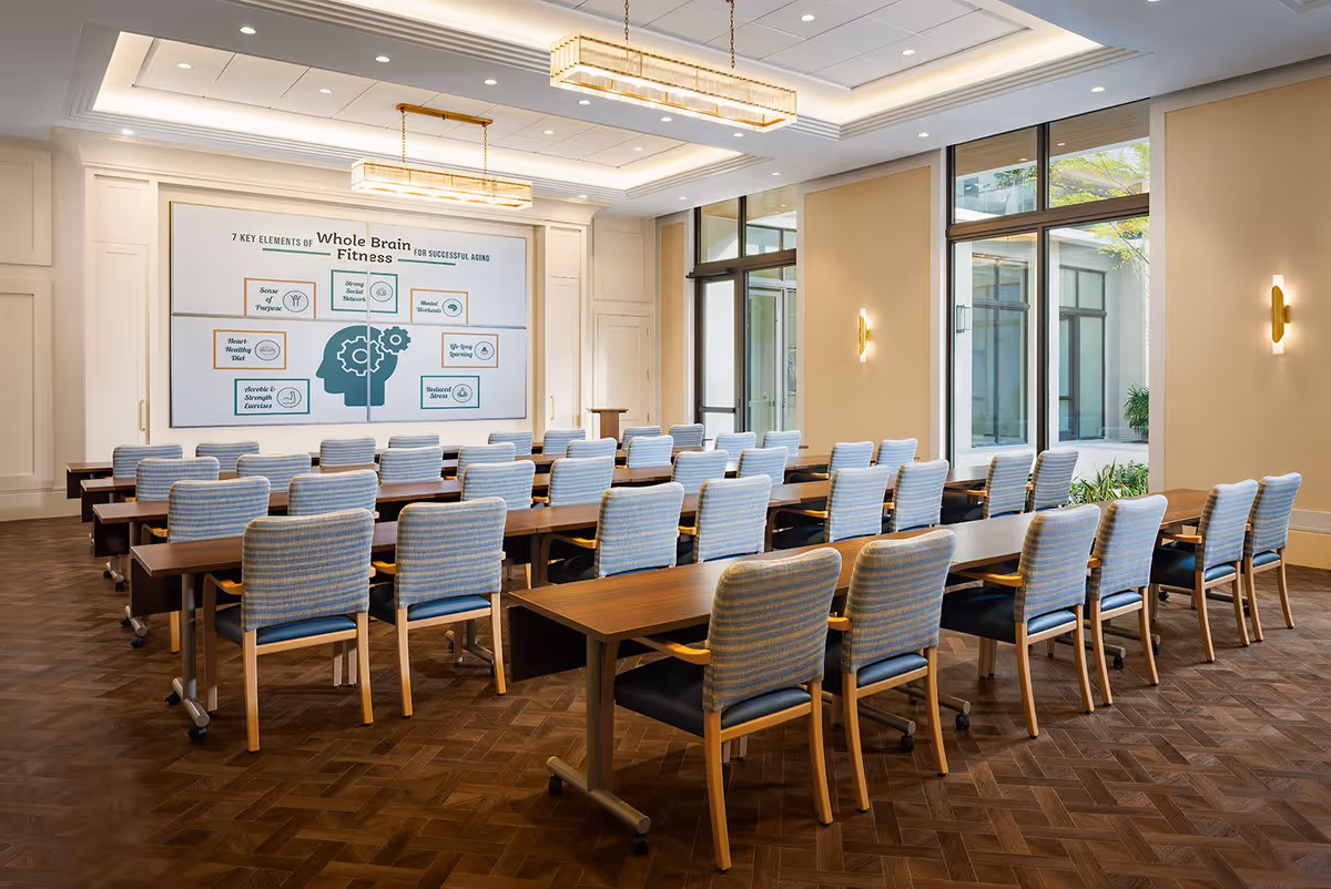 Bright conference-style meeting room with rows of wooden tables and blue upholstered chairs facing a presentation screen.