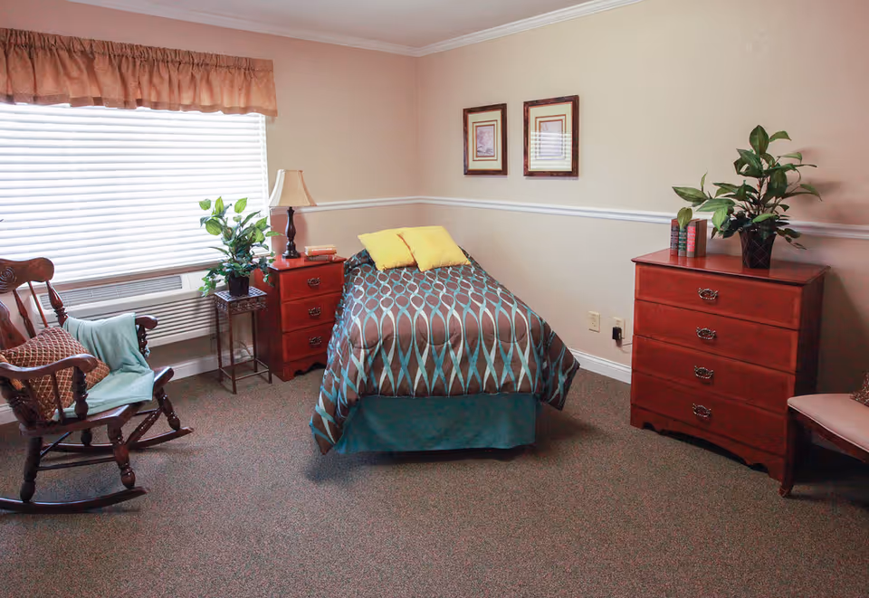 A tidy senior living bedroom with a single bed, red dressers, a rocking chair, and potted plants by a large window.