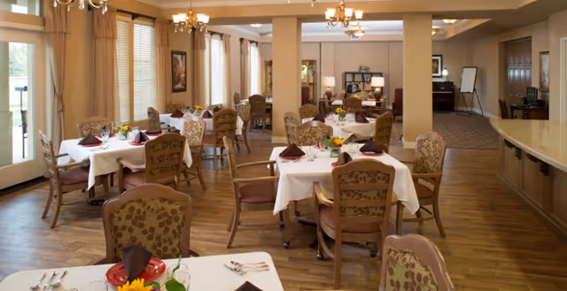 A spacious dining room with multiple tables covered in white tablecloths, each set with plates, silverware, glasses, and folded brown napkins. The room features wooden chairs with patterned upholstery, large windows with beige curtains, and warm lighting from chandeliers. In the background, there is a seating area with sofas and a bookshelf.