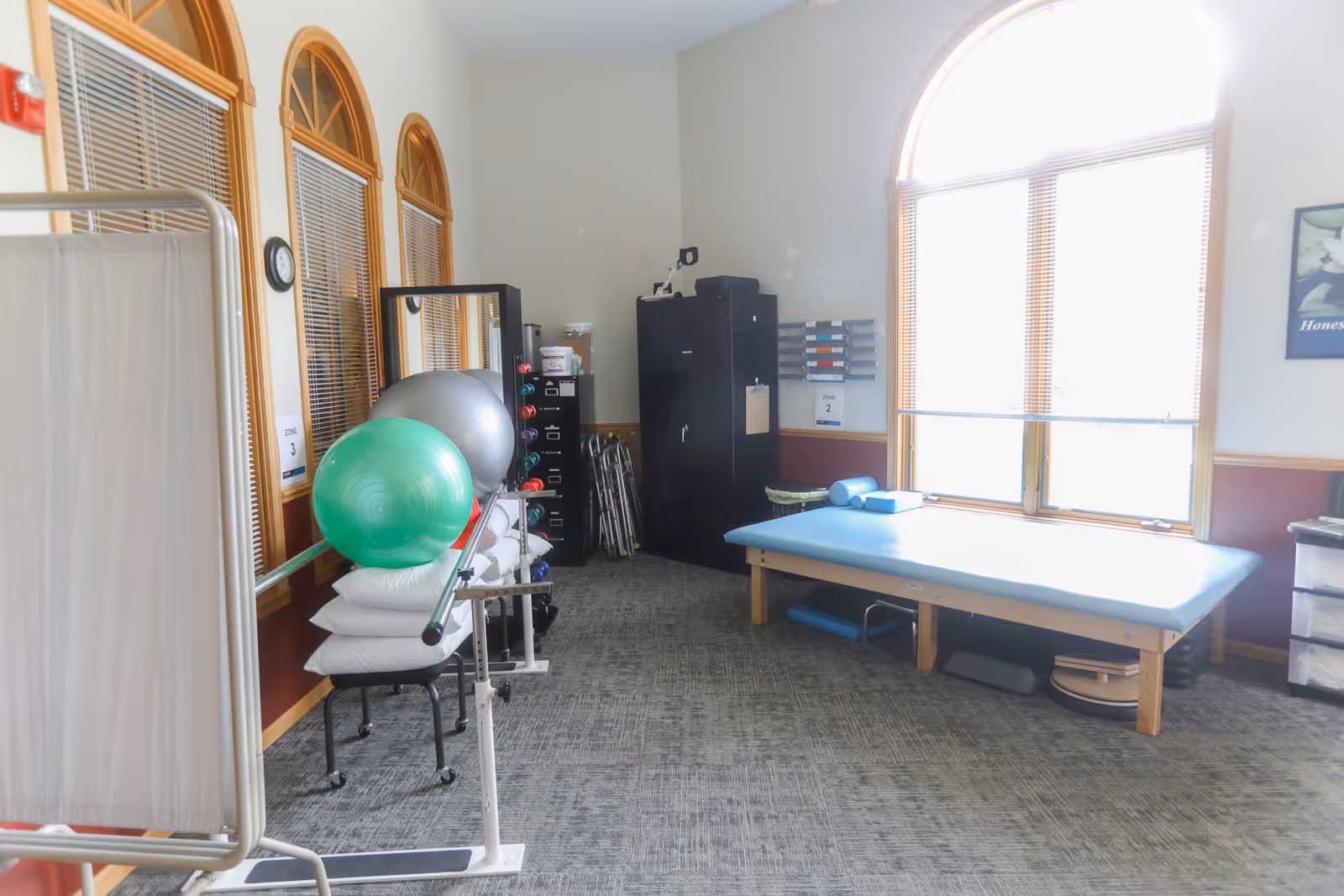 A physical therapy or rehabilitation room with a treatment table near a large window, exercise balls, pillows, weights, and various therapy equipment organized around the room.