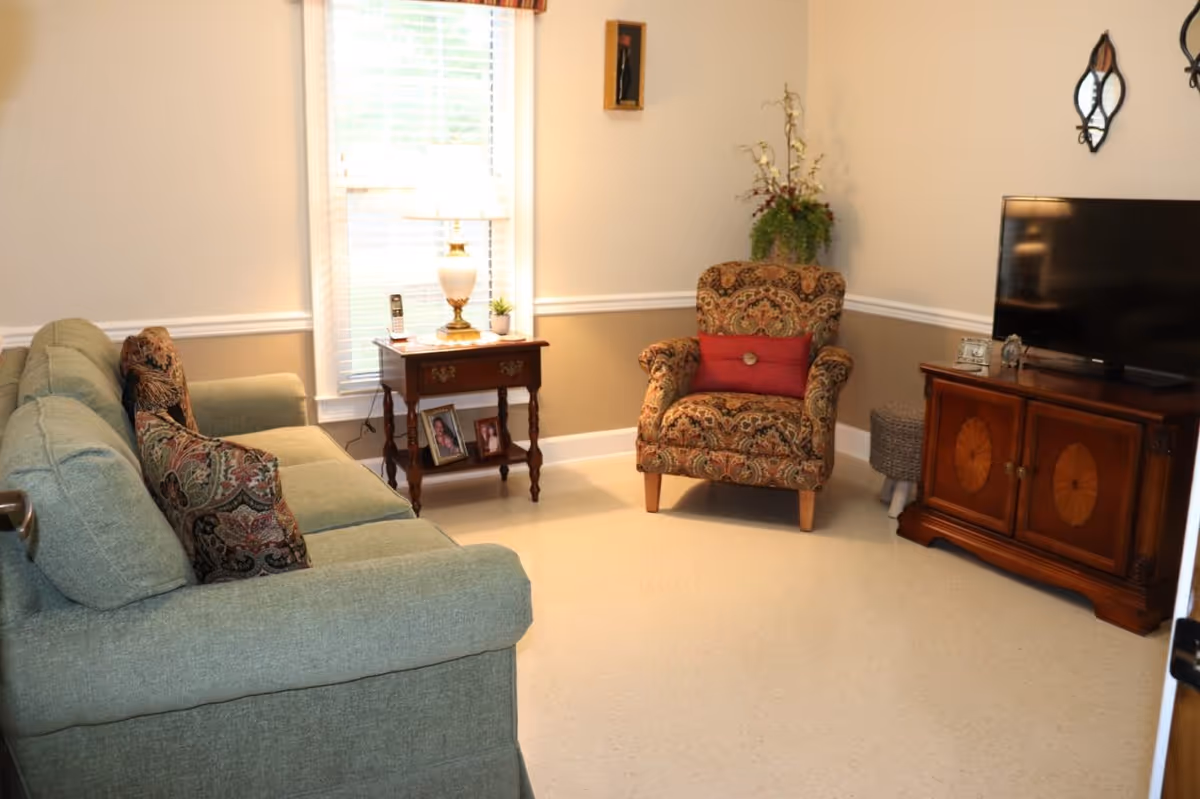A cozy living room with a green sofa adorned with patterned cushions, a patterned armchair with a red pillow, a wooden side table with a lamp and framed photos, a flat-screen TV on a wooden cabinet, and a window with blinds letting in natural light.