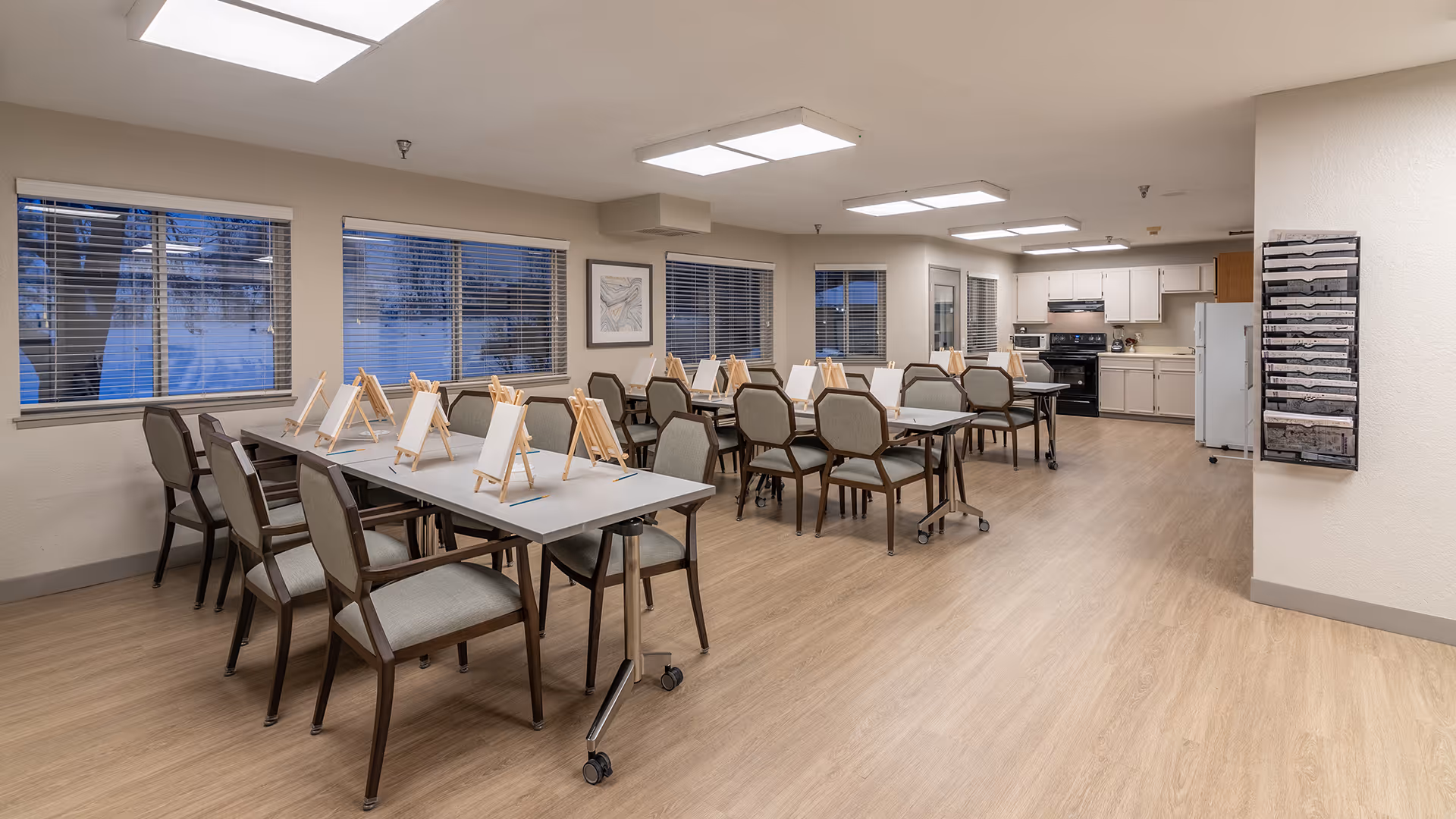 A spacious room with multiple tables and chairs arranged for an art activity, each table has small easels with blank canvases. The room has large windows with blinds, light wood flooring, and a kitchen area with white cabinets, a refrigerator, microwave, and stove in the background.