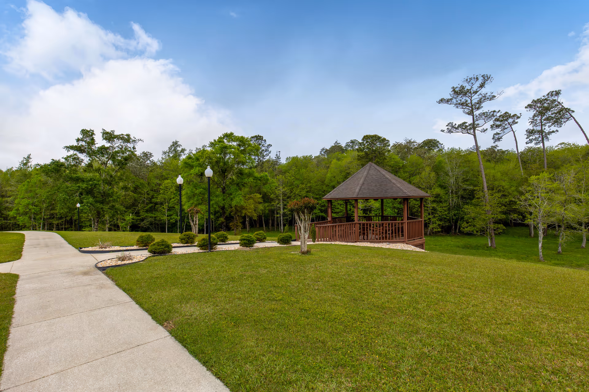 A peaceful outdoor area at Brookside Senior Living Community featuring a paved walkway, neatly trimmed grass, small bushes, lamp posts, and a wooden gazebo surrounded by trees under a partly cloudy blue sky.