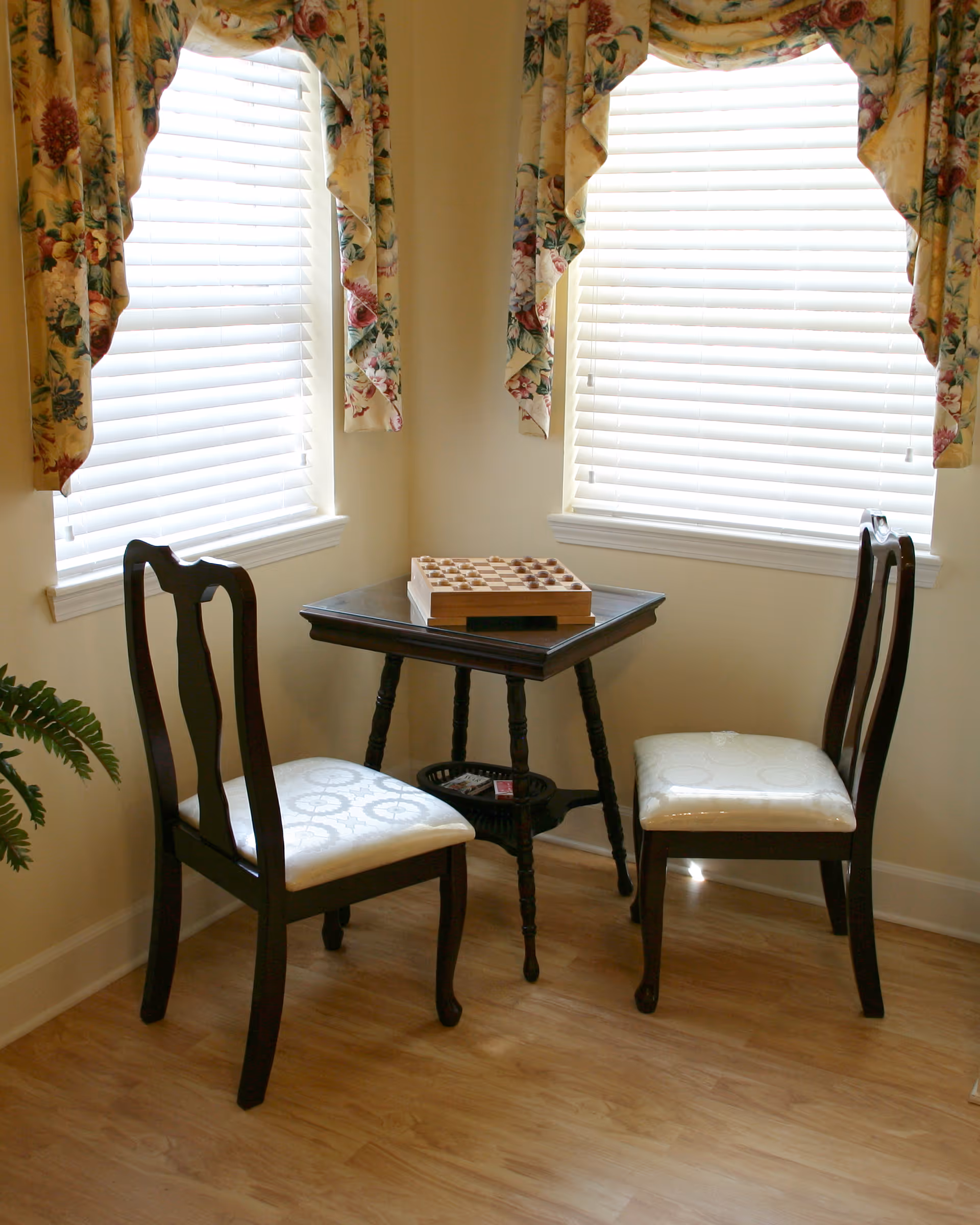 A small corner seating area with two wooden chairs with white cushioned seats facing a small wooden table that has a chessboard on top. The area is illuminated by natural light coming through two windows with white blinds and floral-patterned valances. The floor is light wood, and a green plant is partially visible on the left side.