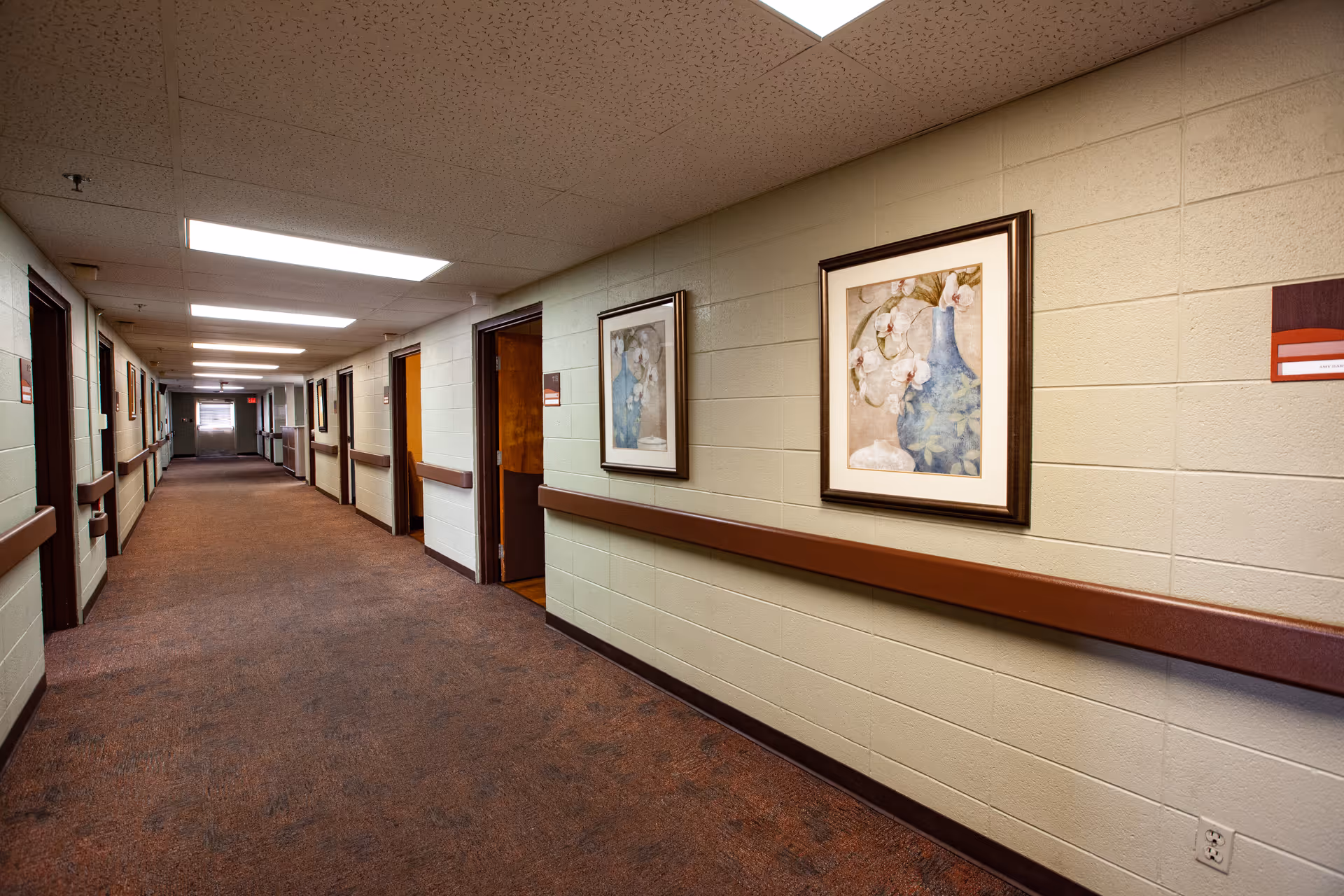 A long hallway in a senior living facility with beige walls, brown handrails, and carpeted floor. Several doorways line the hallway, and framed floral artwork hangs on the walls. The ceiling has fluorescent lighting panels.