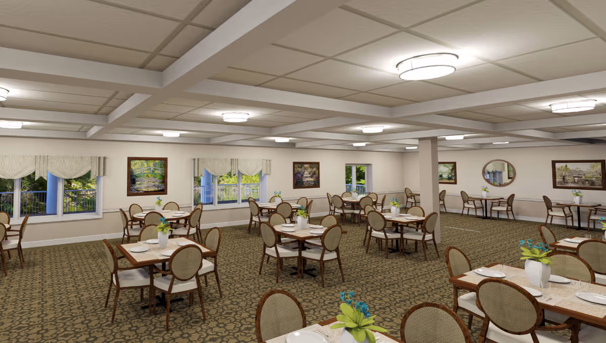 A spacious dining room in a senior living facility with multiple tables and chairs arranged neatly. Each table is set with plates, utensils, and a small vase with flowers. The room has patterned carpet flooring, white walls adorned with framed paintings, and several windows with curtains letting in natural light. The ceiling features recessed lighting and beams.