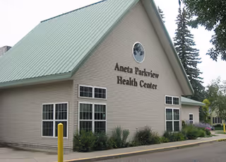 Exterior view of Aneta Parkview Health Center building with a green sloped roof, beige siding, multiple windows, and surrounding greenery including bushes and trees.
