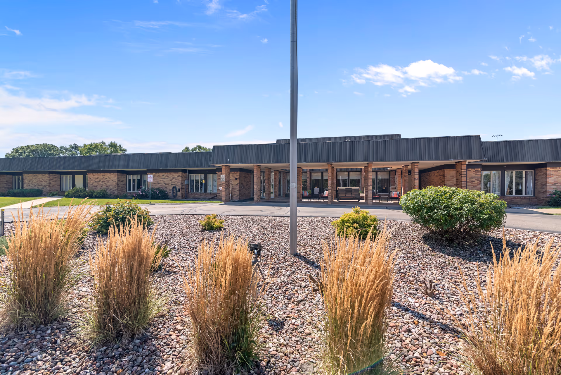 Front exterior view of a single-story brick building with a covered entrance and a landscaped area with ornamental grasses and bushes in the foreground under a blue sky with some clouds.