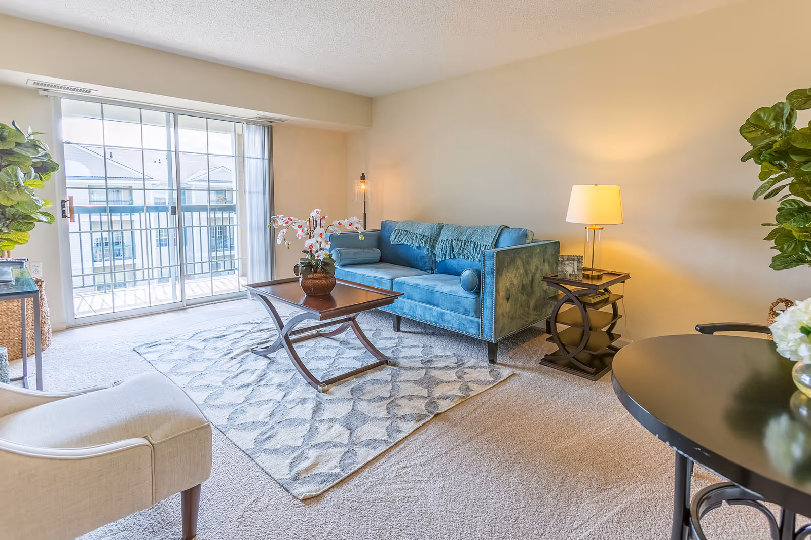 A bright living room with a blue velvet sofa adorned with matching pillows and a throw blanket. In front of the sofa is a wooden coffee table with a decorative flower arrangement. The room has a beige carpet with a patterned area rug. To the right of the sofa is a side table with a lamp and picture frames. Large sliding glass doors lead to a balcony, letting in natural light. There are green plants on either side of the room and a beige armchair partially visible on the left.