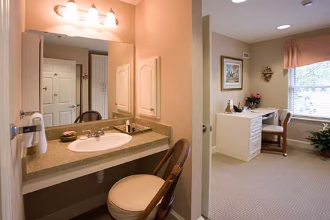 A bathroom vanity area with a sink, countertop, and mirror with three lights above it. A chair is placed in front of the vanity. Adjacent to the bathroom is a room with a desk, chair, framed artwork on the wall, a window with curtains, and a potted plant on the floor.