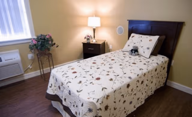 A neatly made single bed with a floral patterned bedspread and matching pillow in a room with wooden flooring. Next to the bed is a dark wooden nightstand with a lamp and a small flower arrangement. There is a window with white curtains, a potted plant on a stand, and a wall-mounted air conditioning unit.