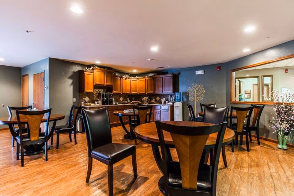 A dining area with several round wooden tables and black chairs with wooden backrests. In the background, there is a kitchen area with wooden cabinets, a coffee machine, and a water cooler. The room has wooden flooring and neutral-colored walls, with a large window on the right side showing an adjacent room.
