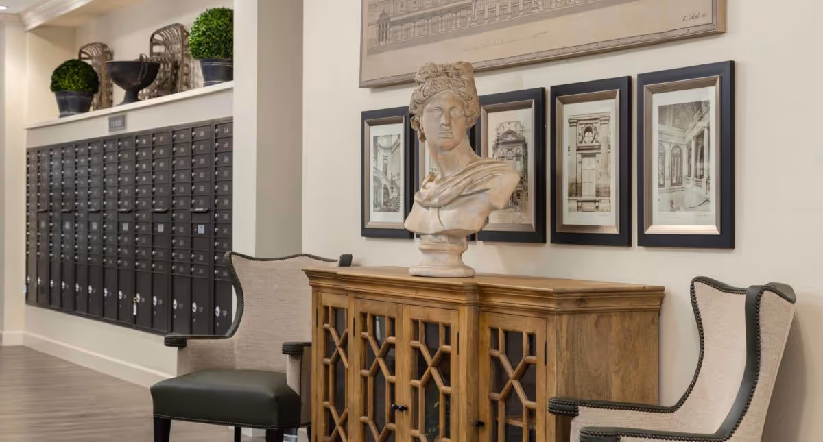 Interior view of a senior living facility mail area with a row of black mailboxes mounted on the wall. In front of the mailboxes, there are two upholstered chairs with dark wooden legs and a wooden cabinet between them. On top of the cabinet is a classical bust sculpture, and above it are framed architectural drawings hanging on the wall.