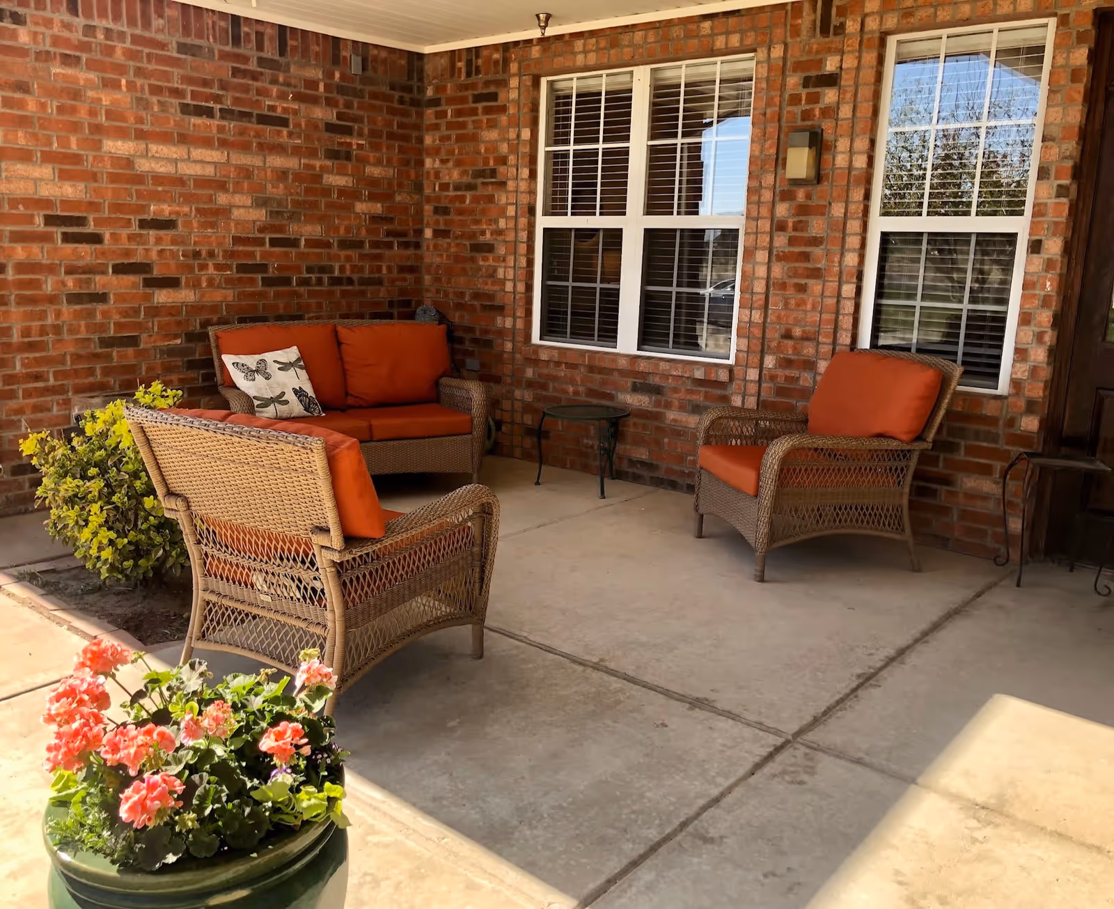 A covered outdoor patio area with brick walls, featuring wicker furniture with orange cushions including a loveseat and two chairs. There is a small glass-top side table between the loveseat and one chair. Two windows with white blinds are visible on the brick wall, along with a wall-mounted light fixture. A green planter with pink flowers is in the foreground.
