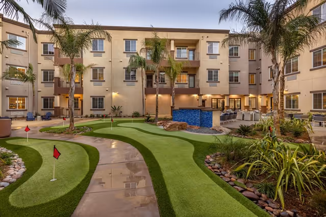 Outdoor courtyard area of a senior living facility featuring a putting green with small red flags, palm trees, landscaped plants, and a seating area with chairs and tables. The building surrounding the courtyard is three stories tall with balconies and windows.