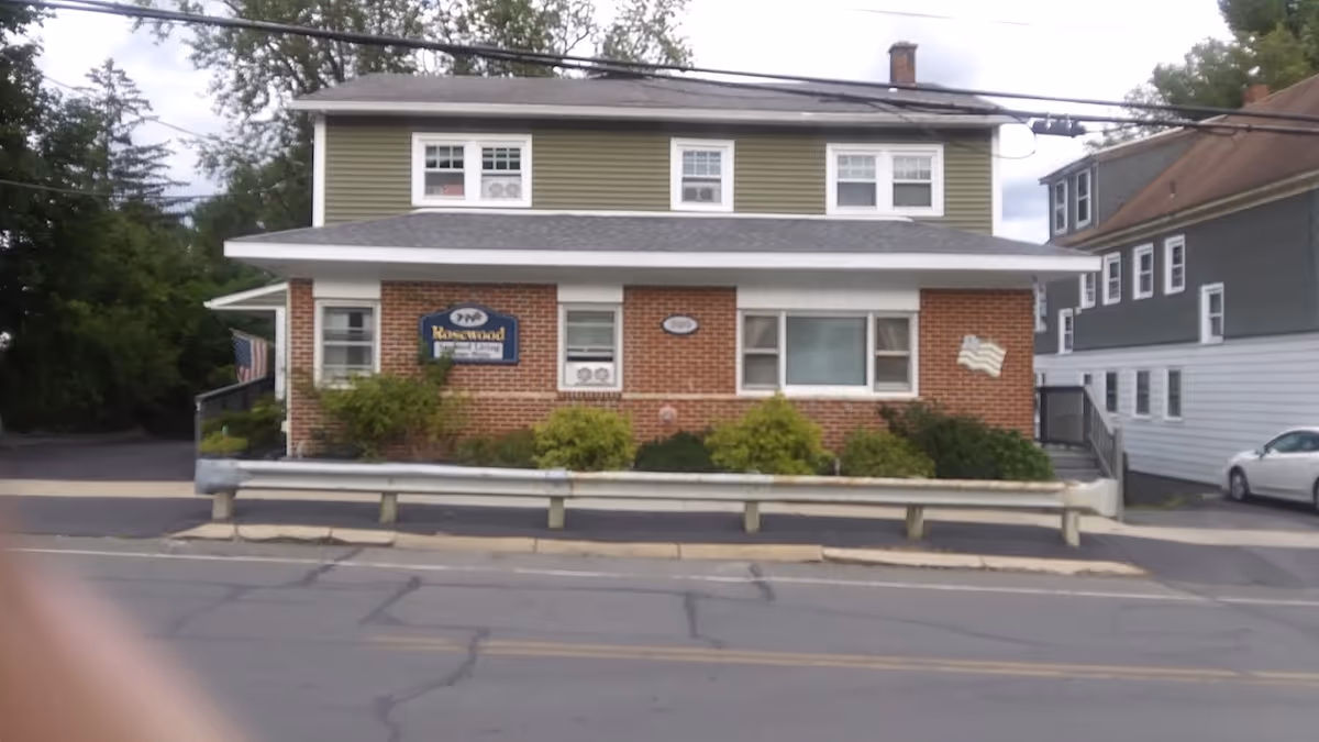 Two-story brick and siding building with a 'Rosewood' sign on the front, shrubs along the foundation and a street with a guardrail in the foreground.