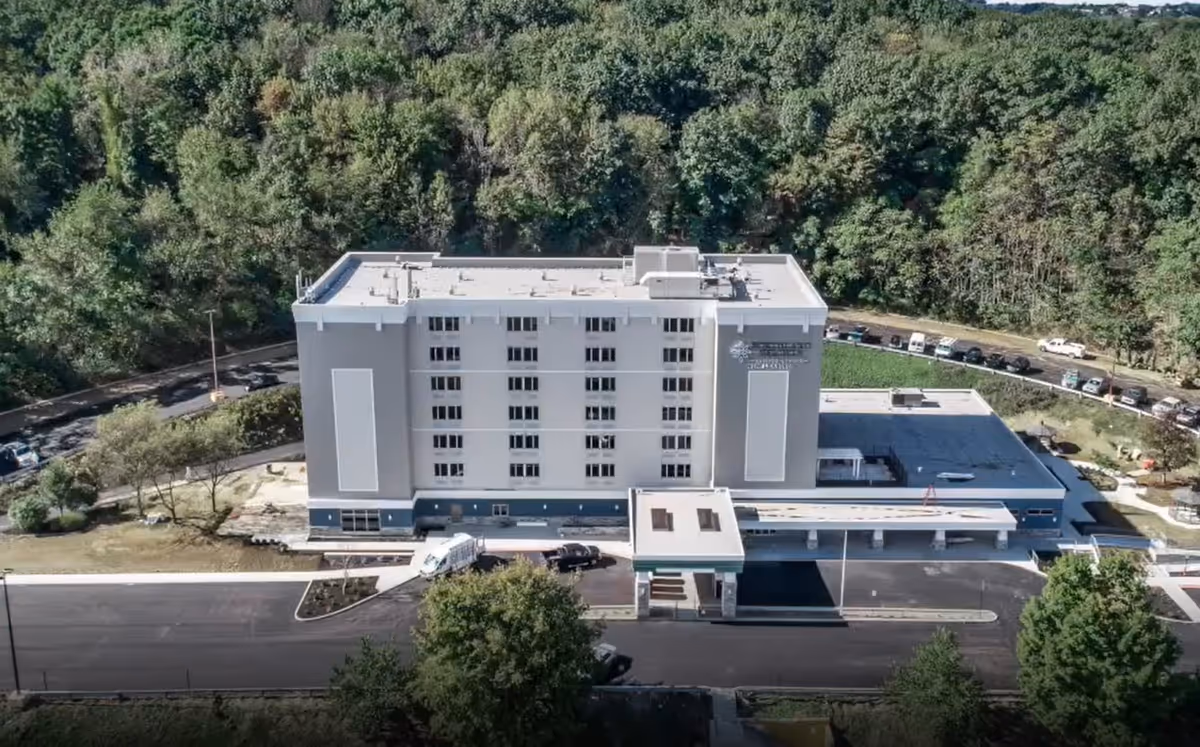 Aerial view of a multi-story apartment building with a covered entrance, parking area, and trees in the background.