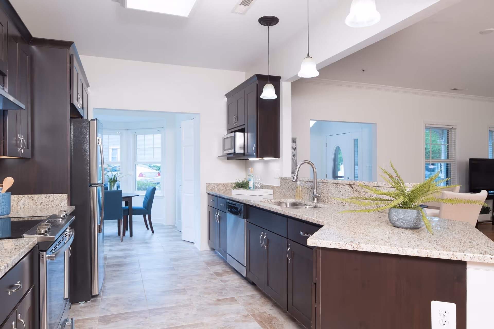 Modern kitchen with dark wood cabinets, granite countertops, stainless steel appliances including a refrigerator, stove, microwave, and dishwasher. There are three pendant lights hanging from the ceiling and a potted fern on the countertop. In the background, a dining area with blue chairs and a round table is visible through an open doorway.