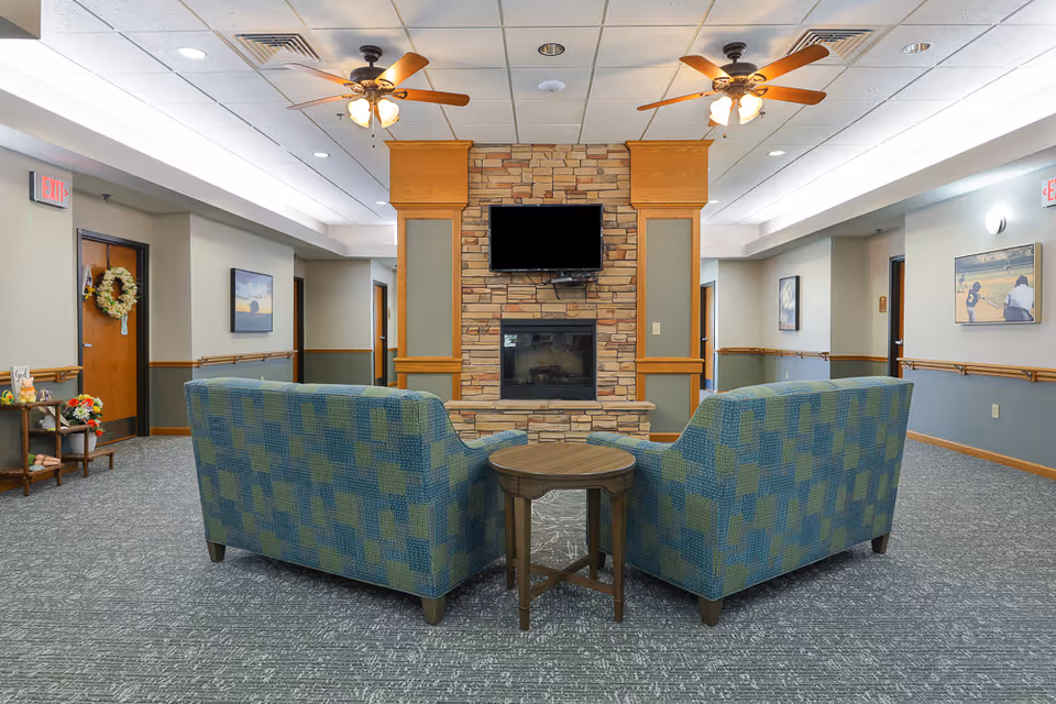 A cozy common area in an assisted living facility featuring two patterned blue and green armchairs facing a stone fireplace with a mounted flat-screen TV above it. The room has a gray carpet, light-colored walls with wooden trim, ceiling fans with lights, and framed artwork on the walls. There are doors and hallways visible on either side of the fireplace.