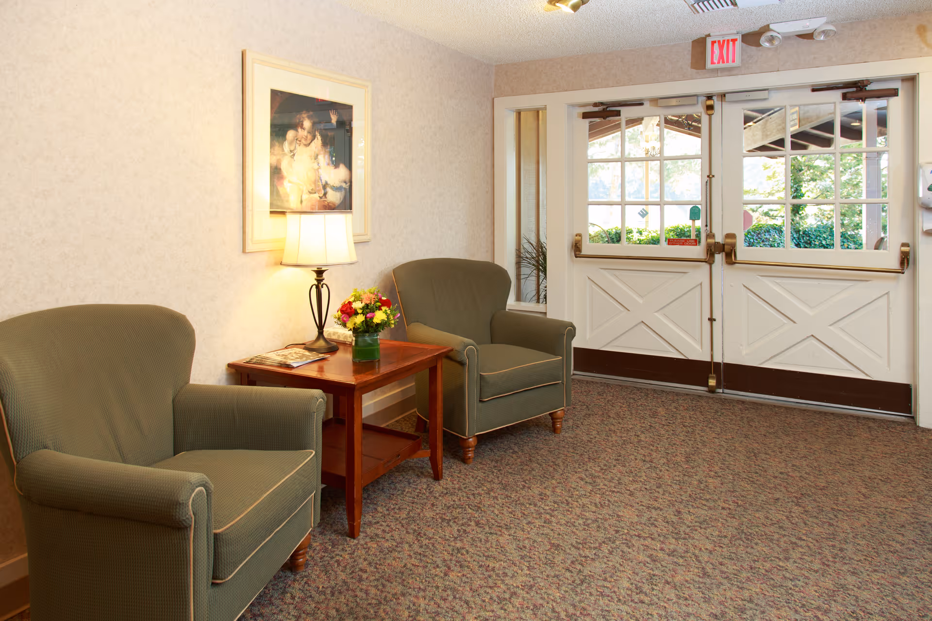 A cozy seating area near an exit door with two green upholstered armchairs and a wooden side table between them. The table holds a lamp and a vase with colorful flowers. A framed picture hangs on the wall above the table. The exit door has glass panels and an illuminated exit sign above it.