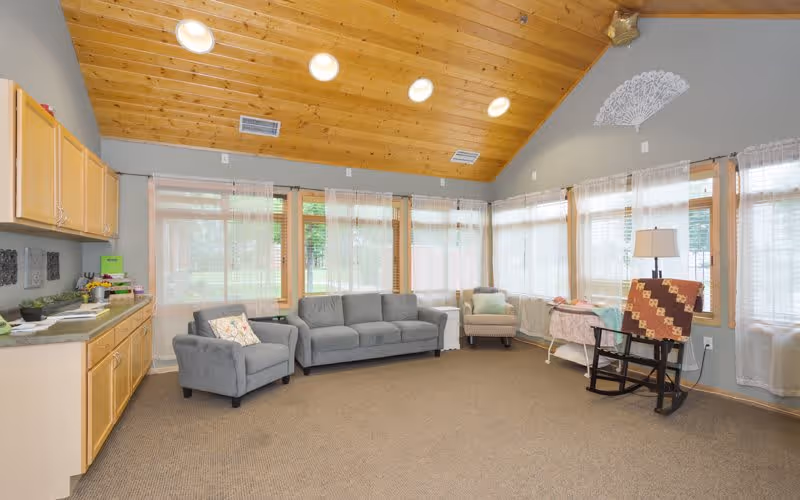 A bright and spacious living room with a high wooden ceiling and multiple large windows covered with sheer white curtains. The room features a gray sofa, two armchairs, a rocking chair with a quilt draped over it, a small side table with a lamp, and a kitchenette area with light wood cabinets and a countertop on the left side.