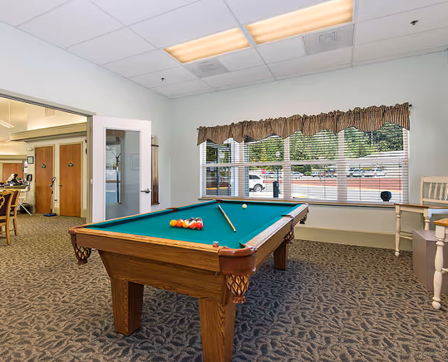 A pool table with cue and racked balls sits in a carpeted common room with large windows and seating.