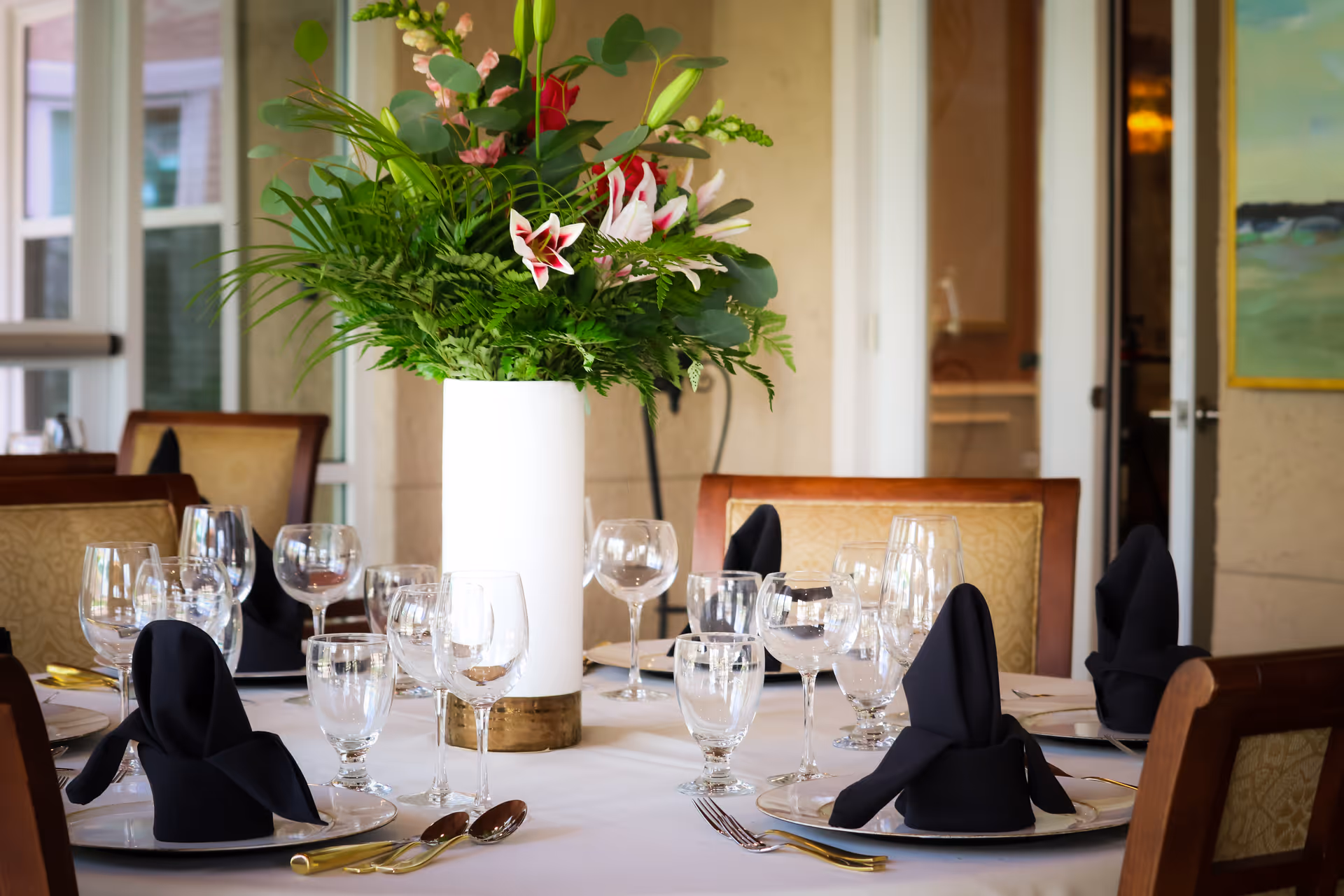 A dining table set with multiple wine glasses, water glasses, plates with black folded napkins, and gold-colored cutlery. A tall white vase with a large floral arrangement featuring green leaves and pink and white flowers is at the center of the table. The background shows upholstered chairs and a door leading to another room.