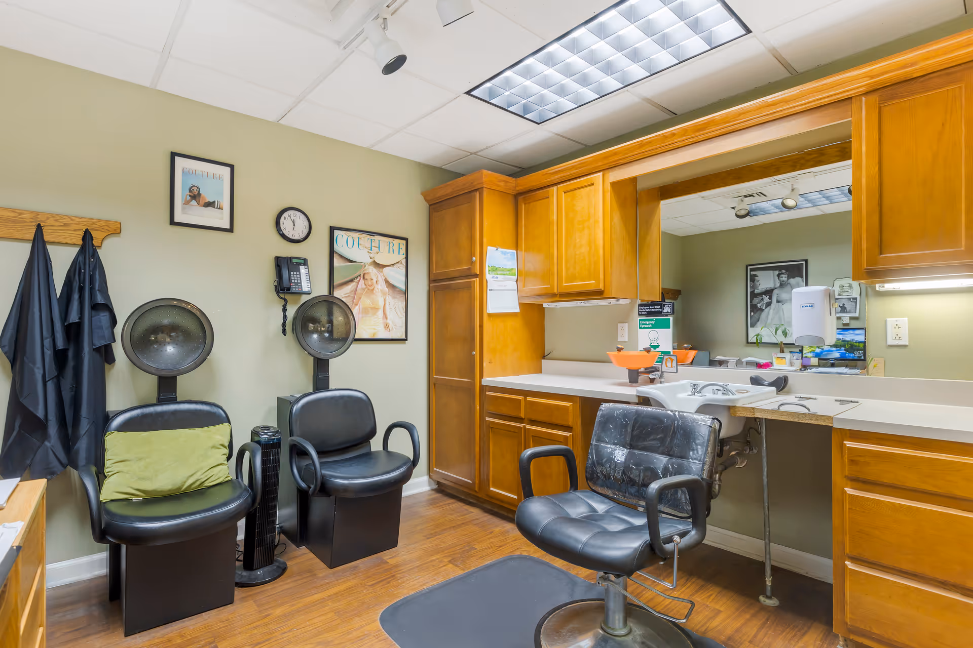 Interior of a salon area in a senior living facility with two black hair drying chairs, a black salon chair in front of a sink, wooden cabinets, a large mirror, and framed pictures on the wall.
