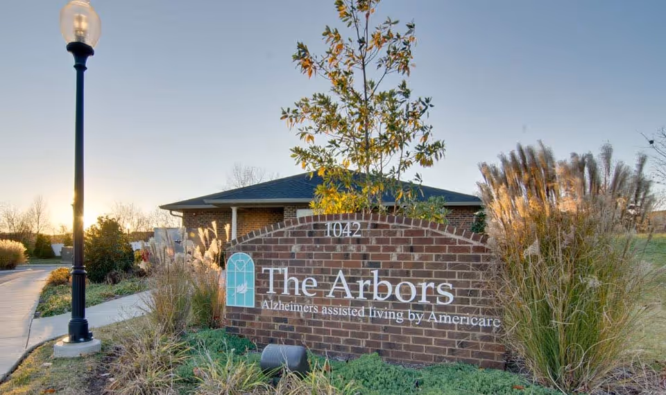 Brick entrance sign reading 'The Arbors Alzheimer's assisted living by Americare' in front of a low brick building with ornamental grasses and a lamppost.