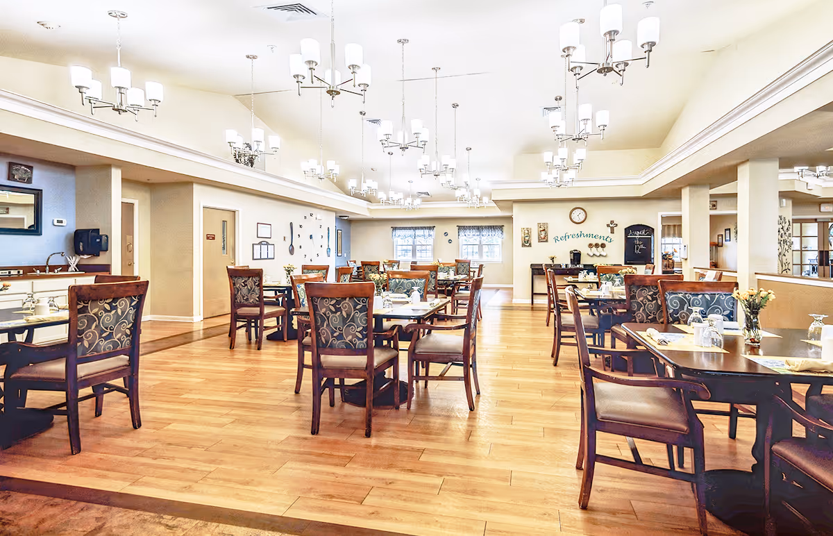 A spacious and well-lit dining room with multiple wooden tables and chairs arranged neatly. The room features light-colored walls, large windows with curtains, and several modern chandeliers hanging from the ceiling. There is a refreshment station against the far wall with a clock above it and decorative wall art around.