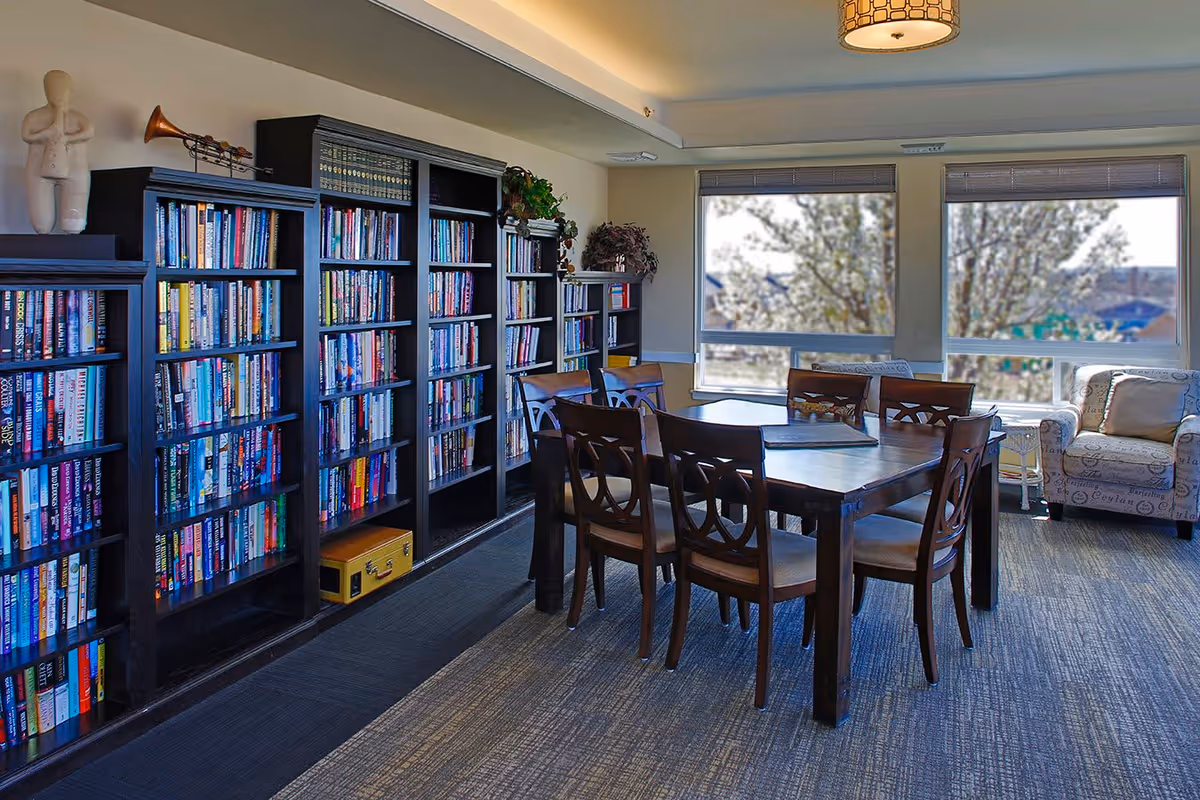 A well-lit room with large windows showing trees outside. The room features several tall bookshelves filled with books along one wall, a wooden table with six chairs in the center, and a cushioned armchair near the windows. The ceiling has recessed lighting and a decorative hanging light fixture.