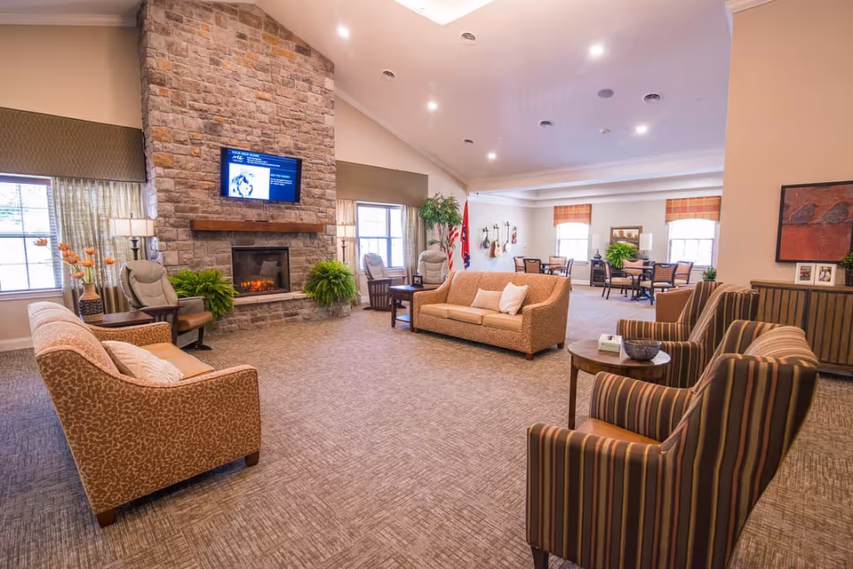 A spacious and well-lit living room area in a senior living facility featuring a large stone fireplace with a mounted TV above it. The room has multiple seating options including patterned sofas, armchairs, and small tables. Large windows with curtains allow natural light to fill the space, and there are several green plants adding a touch of nature. In the background, there are dining tables and chairs near windows with plaid valances.