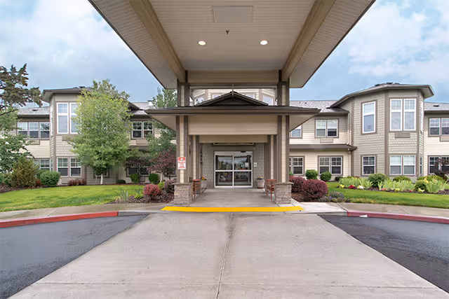 Front exterior view of Brookdale Redmond facility showing a covered entrance with a driveway, landscaped greenery, and a two-story building with multiple windows under a partly cloudy sky.