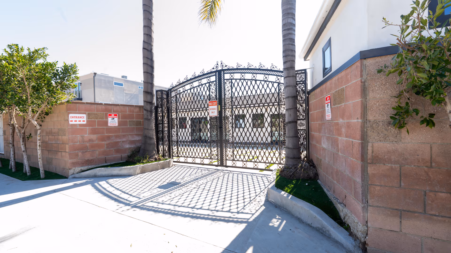 Black wrought iron gate entrance flanked by two palm trees and brick walls with signs indicating entrance and no trespassing. A building is visible behind the gate under a clear sky.