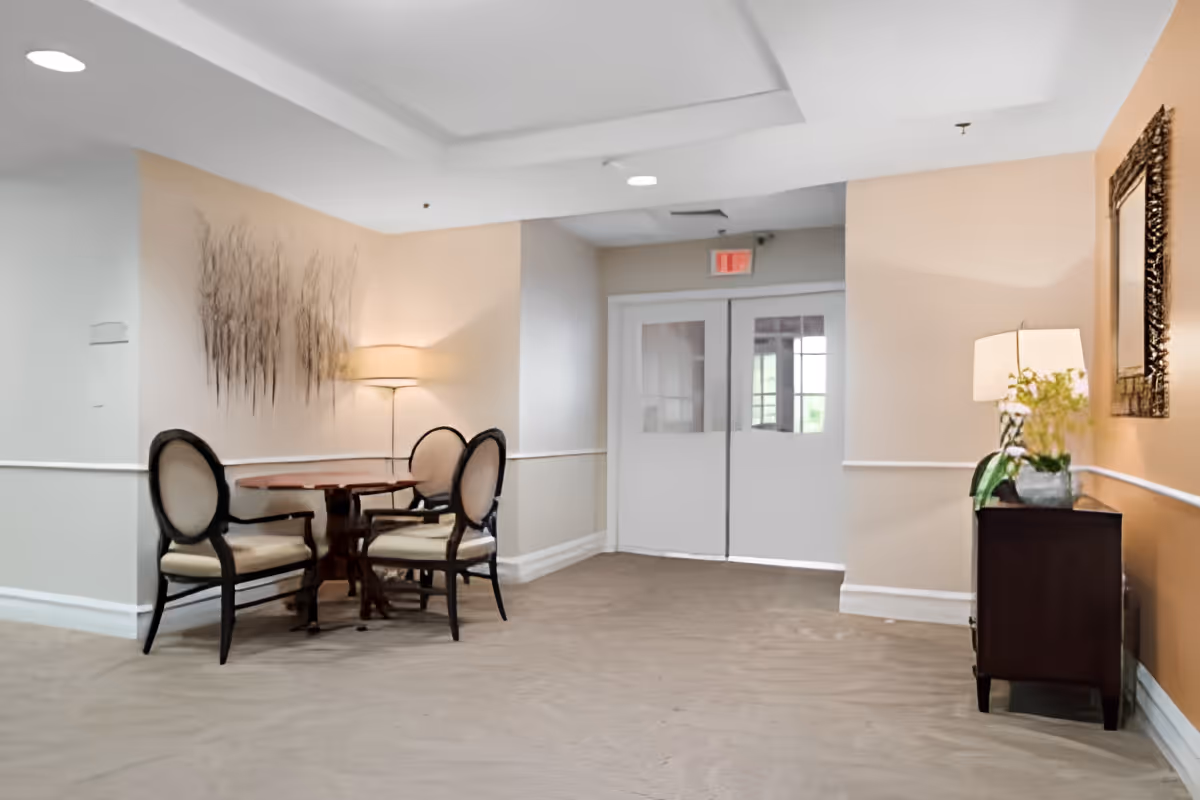 A quiet seating area in a hallway with a round wooden table and three upholstered chairs. The walls are painted in neutral tones with a decorative wall hanging and two lamps providing soft lighting. Double doors with windows are visible at the end of the hallway.
