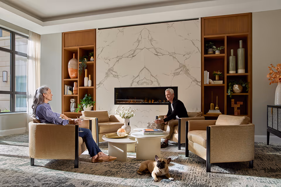 A cozy living room area in The Carnegie at Washingtonian Center featuring two elderly people sitting in beige armchairs around a white coffee table with books and decorative items. A small dog lies on the carpet in front of them. The room has a large window with sheer curtains, built-in wooden shelves with plants and vases, and a modern fireplace set in a marble wall.