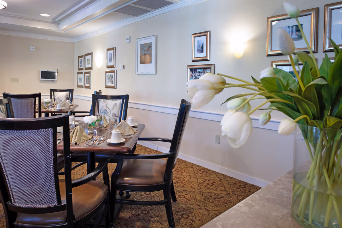 Dining room with a table set for a meal, upholstered chairs, framed wall art, and a vase of white tulips in the foreground.