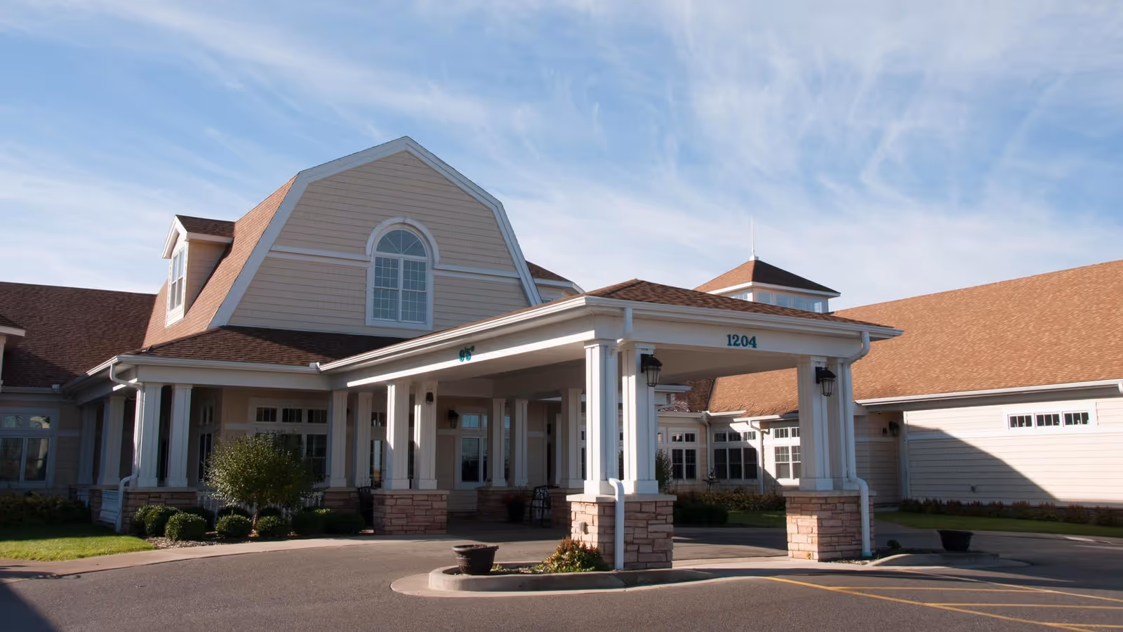 Front entrance and porte-cochère of a single-story senior living facility with columns, a circular driveway, and landscaped beds under a blue sky.