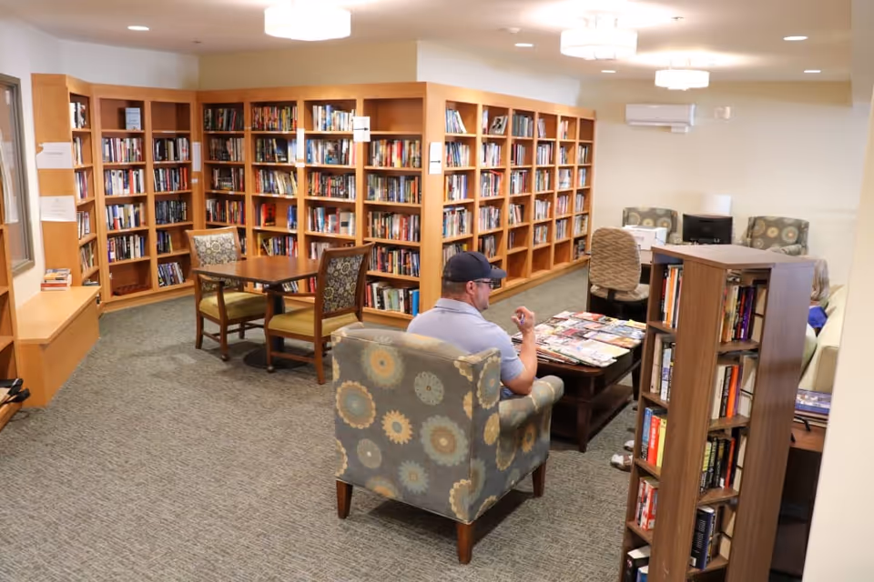 A cozy library lounge with wooden bookshelves, tables, and upholstered chairs and a man seated at a coffee table.