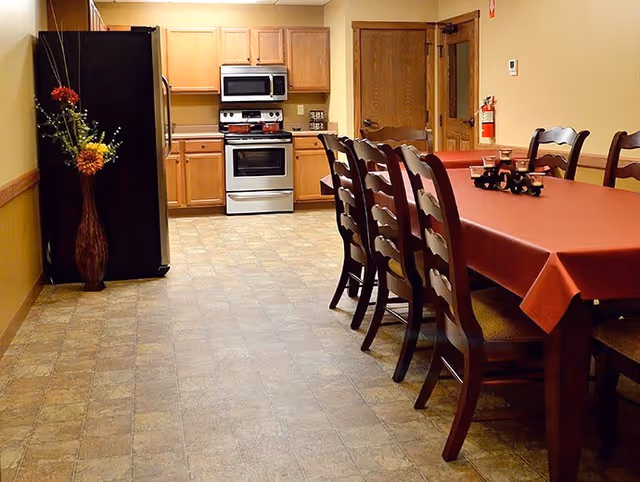 A kitchen and dining area with a long table covered with a red tablecloth and several wooden chairs. The kitchen features wooden cabinets, a black refrigerator, a microwave, and a stove. A vase with flowers is placed near the refrigerator, and a fire extinguisher is mounted on the wall near a wooden door.